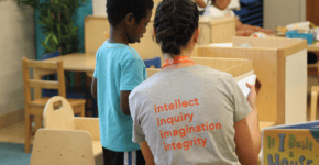 a teacher wearing a gray shirt with orange words that say intellect, inquiry, imagination, and integrity, is working with a young student.