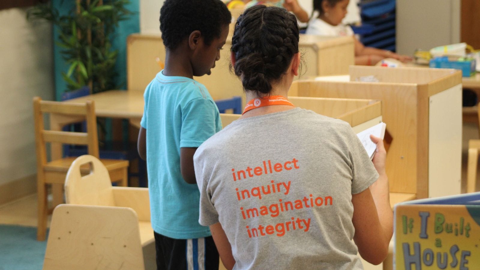 a teacher wearing a gray shirt with orange words that say intellect, inquiry, imagination, and integrity, is working with a young student.